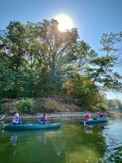 Three people kayak on a calm river surrounded by lush trees under a bright sun.