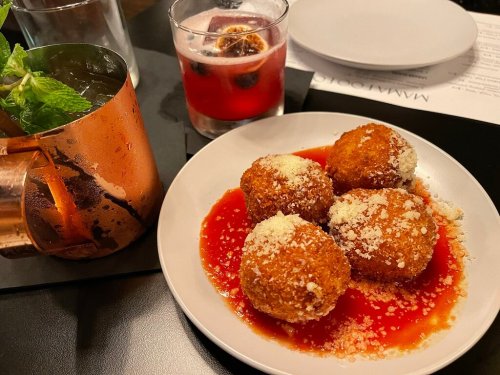 A plate of golden-brown arancini balls drizzled with red sauce and sprinkled with cheese, alongside two colorful cocktails.