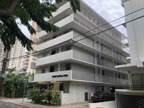 A modern, multi-story hotel building with balconies, surrounded by greenery and urban architecture.