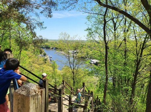 A scenic view of a river surrounded by lush greenery, with people enjoying the outdoors on a sunny day.
