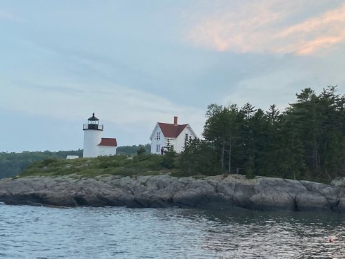 A white lighthouse and a house on a rocky shore, surrounded by trees and water under a cloudy sky.