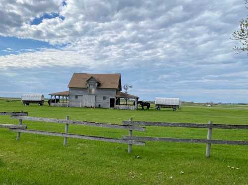 A white farmhouse with a brown roof sits in a green field, surrounded by wagons and under a cloudy sky.