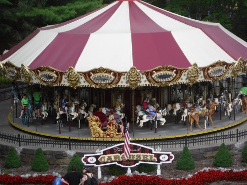 A colorful carousel with ornate decorations, featuring horses and riders, surrounded by flowers and greenery.