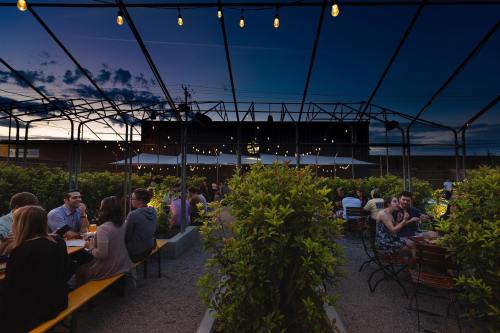 A lively outdoor dining area with string lights, surrounded by greenery, as people enjoy their meals at dusk.