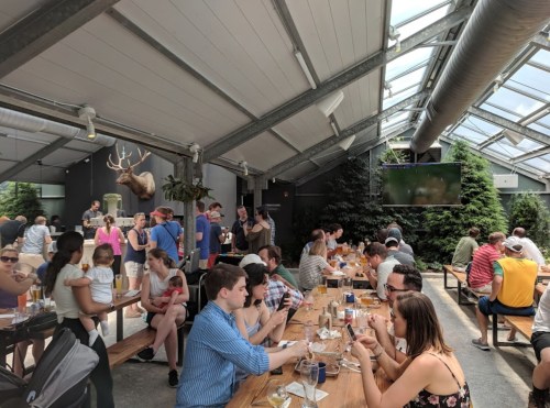 A lively indoor space with people dining and socializing at long tables, surrounded by greenery and a mounted deer head.