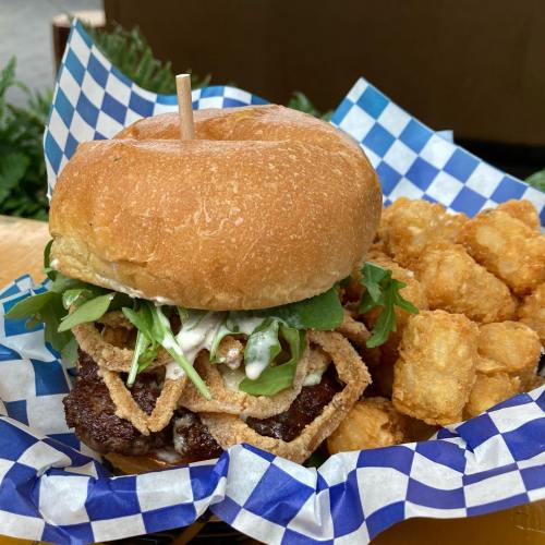 A burger with lettuce, sauce, and crispy onions, served with a side of golden tater tots on a blue checkered plate.