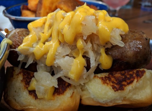 A close-up of a sausage topped with mustard and sauerkraut, served on a toasted bun with golden-brown potato wedges.