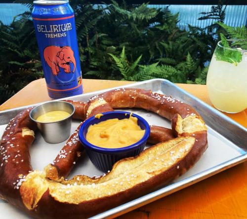 A large pretzel with mustard and cheese dip, alongside a can of Delirium Tremens beer and a cocktail on a tray.
