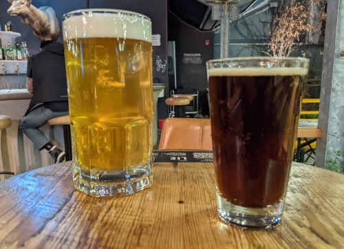 Two glasses of beer on a wooden table: a tall, light-colored beer and a shorter, dark beer.