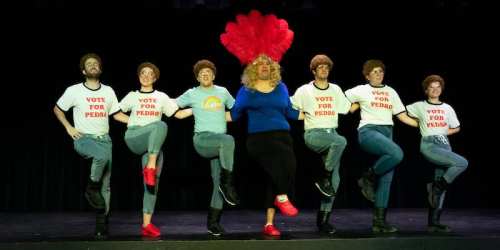 A group of performers in matching shirts and wigs, dancing with a central figure in a blue top and blonde wig.