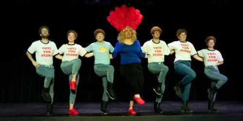 A group of performers in matching shirts and wigs, dancing with a central figure in a blue top and blonde wig.