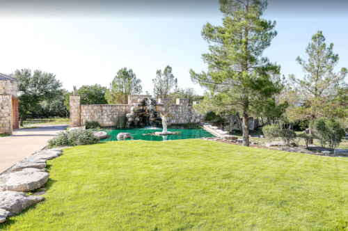 A lush green lawn with a stone fountain and trees, surrounded by a serene pool and landscaped garden.