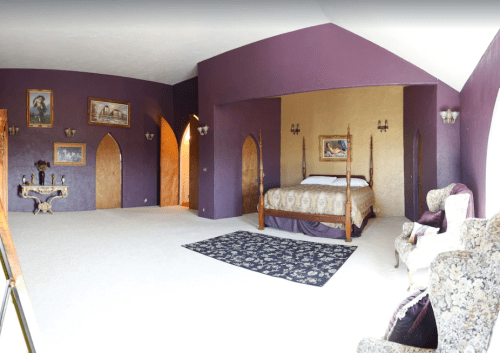 A spacious bedroom with purple walls, a four-poster bed, elegant furniture, and decorative artwork.