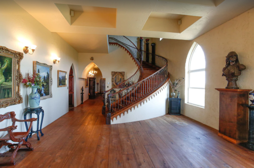 A spacious hallway with a curved staircase, wooden floors, and ornate artwork on the walls. Natural light from large windows.