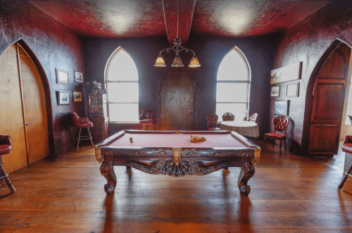 A vintage-style room with a pool table, ornate decor, and large arched windows, featuring dark walls and wooden flooring.