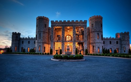A grand stone castle with turrets, illuminated at dusk, surrounded by manicured gardens and a circular driveway.