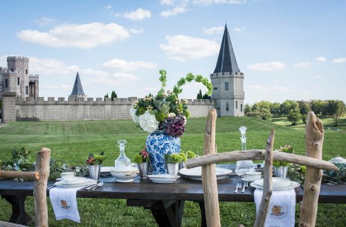 A beautifully set outdoor dining table with floral arrangements, overlooking a castle and green landscape under a blue sky.