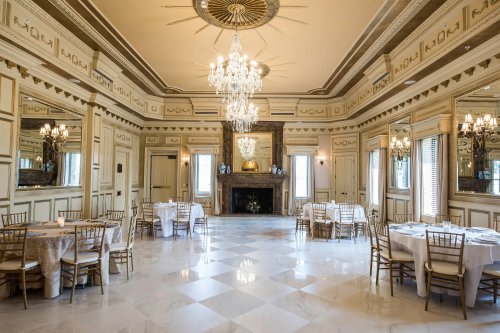 Elegant banquet hall with chandeliers, marble floors, and tables set for dining, featuring ornate mirrors and a fireplace.