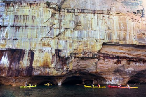 Kayakers explore a scenic coastline with towering cliffs and caves reflected in calm waters.