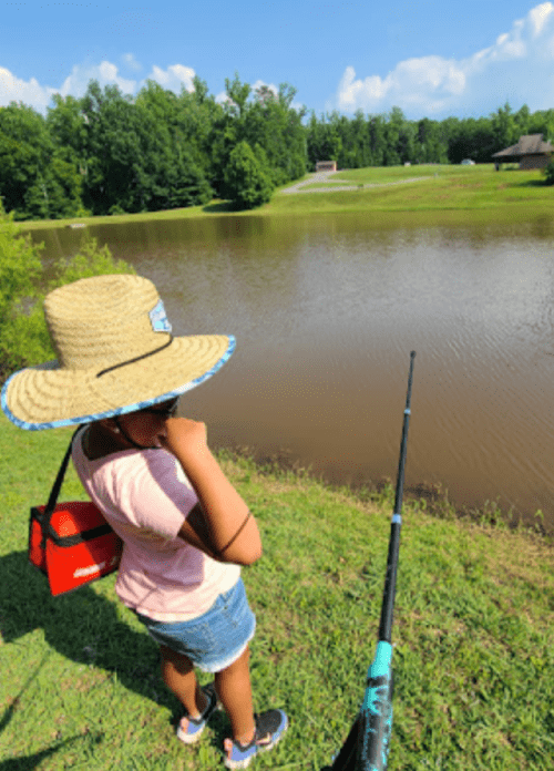 A child wearing a large straw hat stands by a pond, holding a fishing rod, with trees and a building in the background.