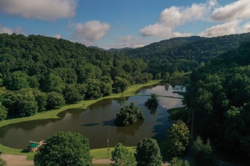 Aerial view of a serene lake surrounded by lush green trees and rolling hills under a partly cloudy sky.