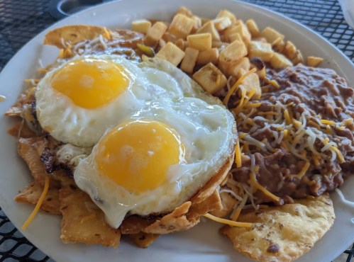 A plate of nachos topped with two fried eggs, cheese, and a side of diced potatoes and refried beans.