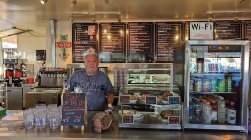 A smiling man stands behind a counter at a café, with a menu board and various food items displayed.