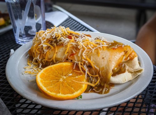 A plate with a large burrito topped with cheese and sauce, accompanied by a slice of orange.