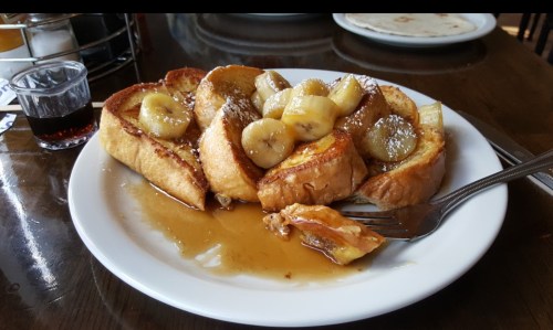 A plate of French toast topped with sliced bananas and syrup, served with a fork on the side.