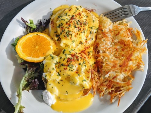 A plate of eggs Benedict topped with hollandaise sauce, served with hash browns and a slice of orange.