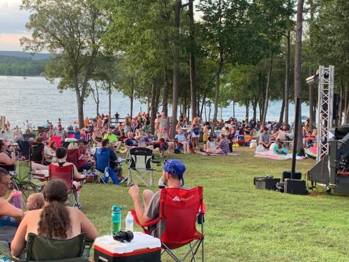 A large crowd gathers on a grassy area by a lake, with people sitting in chairs and enjoying the outdoors.