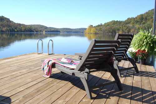 Two lounge chairs on a wooden dock overlooking a calm lake surrounded by autumn foliage.