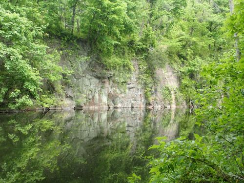 A serene river scene with lush green trees and a rocky cliff reflected in calm water.