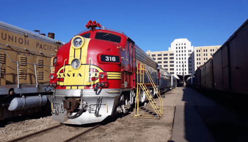 A vintage red and yellow train engine parked beside a row of freight cars, with a city skyline in the background.