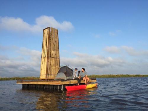 Two people sit on a kayak near a wooden structure on a floating platform in a calm body of water under a blue sky.