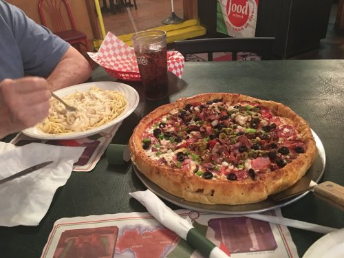 A table with a large pizza topped with meats and vegetables, alongside a plate of pasta and a drink.
