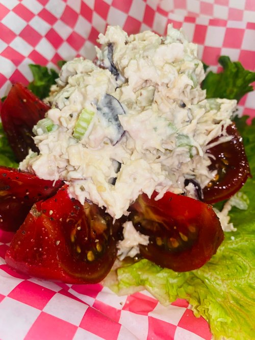 A serving of chicken salad on a bed of lettuce, topped with sliced tomatoes, in a red and white checkered basket.