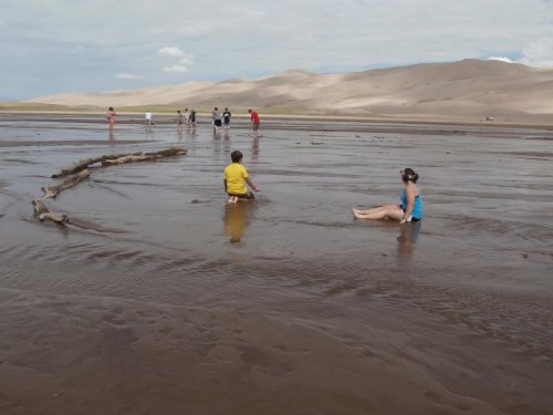 Children playing in shallow water at a sandy beach, with hills in the background and others exploring nearby.