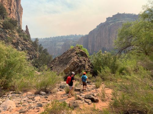 Two hikers walk along a rocky trail surrounded by lush vegetation and towering cliffs under a hazy sky.