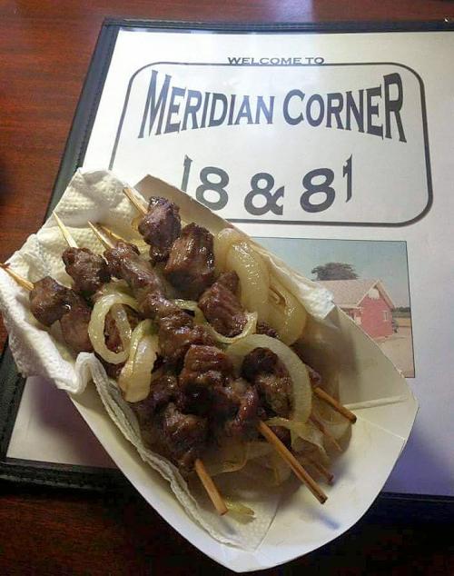 A plate of skewered meat and onions served on a paper tray, with a sign for Meridian Corner in the background.