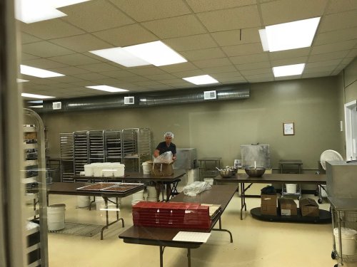 A baker prepares ingredients in a spacious kitchen with tables, racks, and baking supplies.