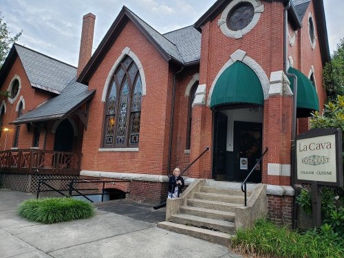 A brick building with arched windows, featuring a sign for La Cava Italian Cuisine and a person sitting on the steps.