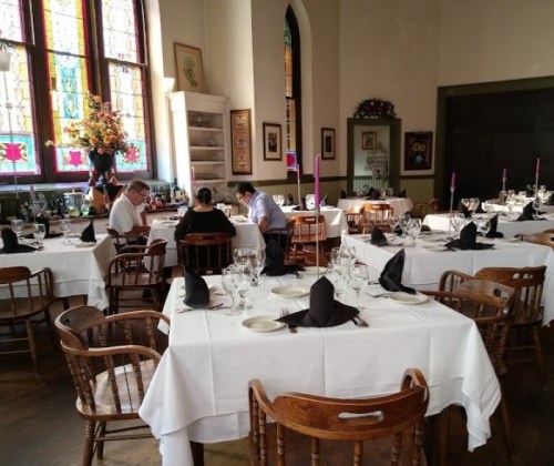 A cozy restaurant interior with wooden chairs, white tablecloths, and stained glass windows, set for dining.