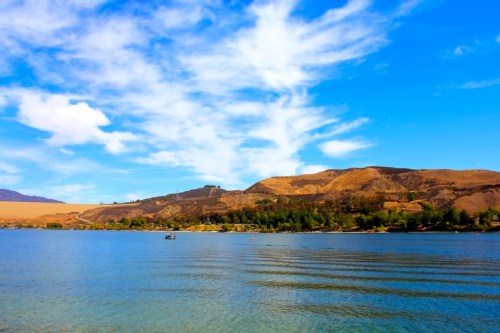 A serene lake surrounded by rolling hills and a clear blue sky with scattered clouds.