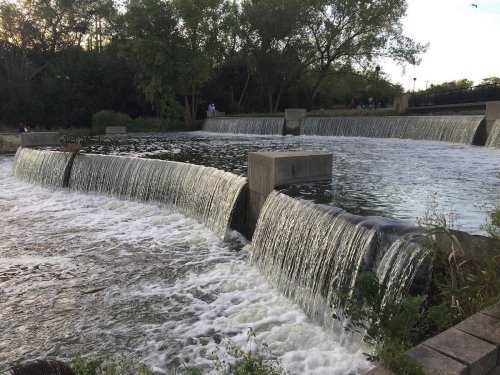 A serene waterfall cascades over stone barriers, surrounded by lush greenery and a calm river.