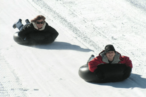 Two people sliding down a snowy hill on black inner tubes, enjoying a winter day.