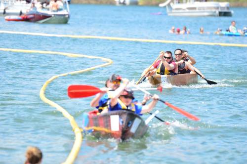 Teams compete in a cardboard boat race on a sunny day, paddling vigorously in a lake surrounded by spectators.