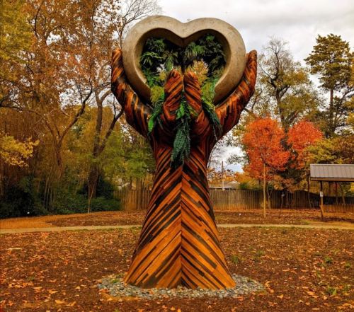 A large wooden sculpture of hands holding a heart, surrounded by colorful autumn trees in a park.