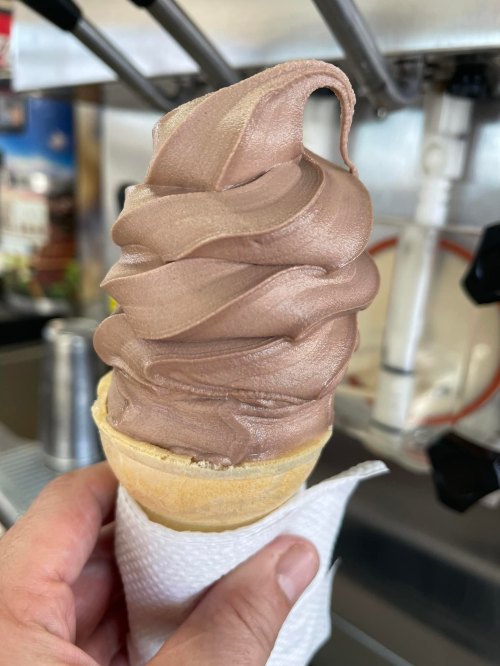 A hand holding a chocolate soft serve ice cream cone with a swirl on top, wrapped in a napkin.