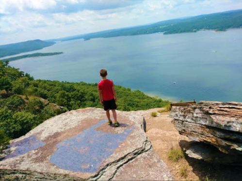 A child stands on a rocky ledge overlooking a scenic lake surrounded by green hills under a blue sky.